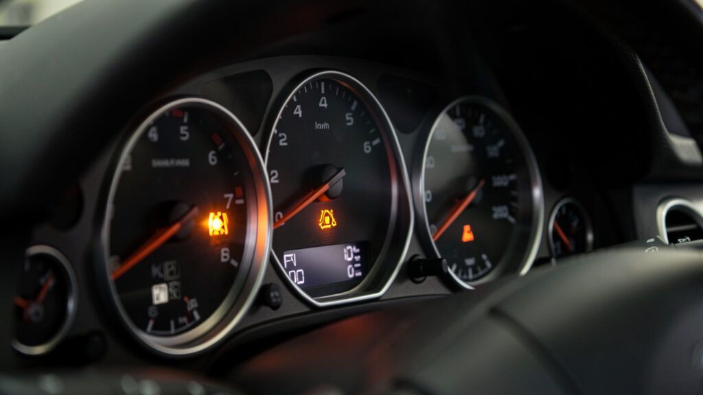 Gros plan réaliste du tableau de bord d’une voiture avec un voyant orange d’injection allumé, éclairage technique et détails des cadrans visibles.