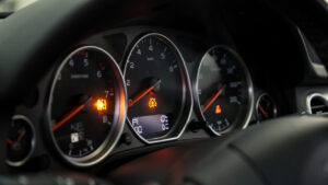 Gros plan réaliste du tableau de bord d’une voiture avec un voyant orange d’injection allumé, éclairage technique et détails des cadrans visibles.