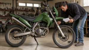 Un homme inspecte le moteur d’une moto d’aventure verte d’occasion dans un garage lumineux, avec des documents d’entretien vierges à la main.